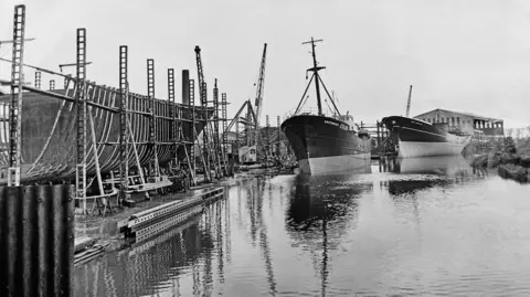 Mirrorpix via Getty Images A view looking down a river with a shipyard on the left. Two trawlers are moored in the river, while the hull of a third is being built. A crane and a warehouse can also be seen.