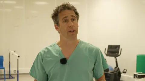 Dr David Hutchins - a man with short dark brown curly hair and with brown eyes. He is wearing green hospital scrubs and is stood in a gym.