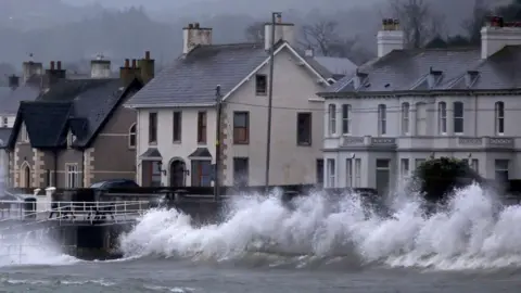 Large waves crash against a sea wall. Behind them is a row of houses. 