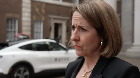 Liz Kendall wearing a serious expression, dressed in a dark suit, stands in front of a white car