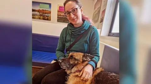 A woman wearing black leggings and a blue hoodie, with glasses and her hair in a ponytail. She's sitting on a blue bench with a German Shepherd dog lying next to her with its head on her lap, and she has her arms around it. They're in a room with white walls, with posters and a window behind her.