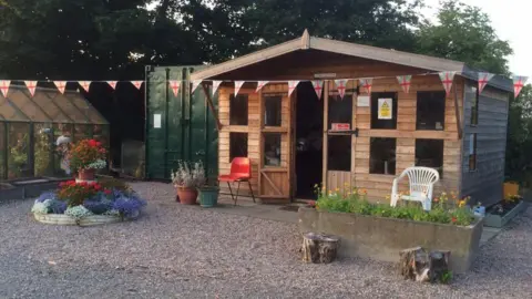 A cabin on an allotment which has flowers and plants outside and a few garden chairs. There are two greenhouses next to it and ENgland flag bunting across the top.