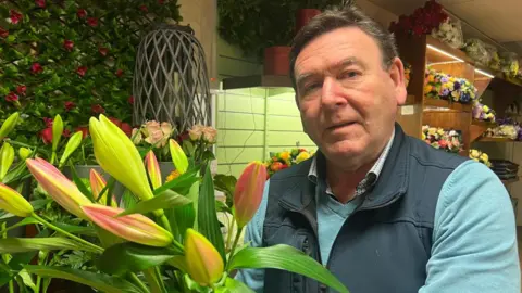 Michael O'Neill is holding a large bouquet of flowers. He is wearing a navy gilet and green fleece. 