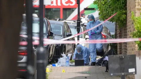 PA Media Forensic officers in blue protective suits examine evidence on a pavement cordoned off with police tape. Numbered evidence markers and investigation materials are visible beside parked cars and a brick wall.