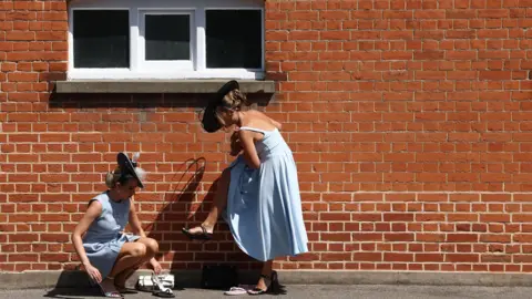 Reuters Two women, dressed in blue dresses, stand next to the brick wall of a building. One on the right hand side is standing up, with a foot in the air as if she is trying to fix a buckle or tie on it. The other on the left side is kneeling down, looking like she is putting on a shoe or helping the other woman.
