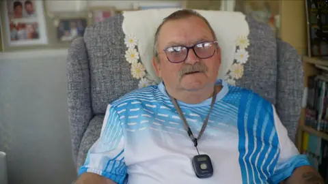 Jamie Niblock/BBC A man with short grey hair is wearing glasses and a blue and white-striped T-shirt and is sitting in a grey-coloured armchair. He has a small rectangle-shaped dark grey fall pendant around his neck. 