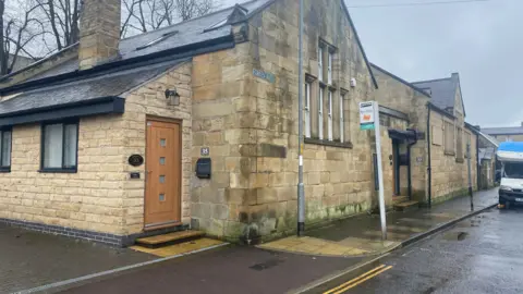 A Victorian building which was once a school with high windows has two gables either side of a main door. A separate more modern entrance is at the forefront of the building