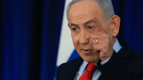 Benjamin Netanyahu stands speaking at a press conference. He wears a black suit jacket, blue shirt and red tie. One of his hands is raised, pointing his finger and he speaks. In the background, the Israeli flag is blurred.