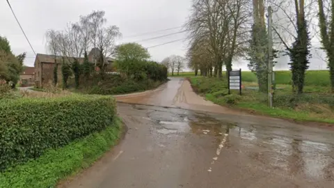 Google A rural crossroads surrounded by fields, trees and hedgerows and a large building to the left.