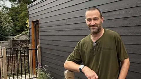 Hopestead A smiling man with a green T-shirt and black trousers standing in front of a black and wooden pod for the homeless where he now lives