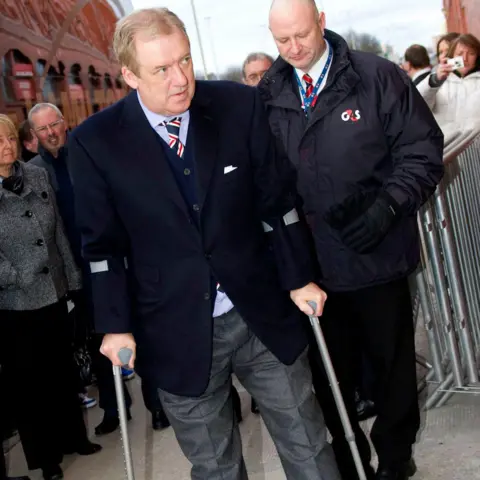 SNS Sir David Murray outside Ibrox in 2011. He is wearing a dark blazer, a light blue shirt and a red and blue striped tie. He walks on crutches. A security guard stands behind him.