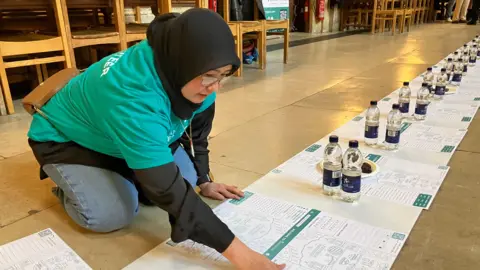 Shariqua Ahmed/BBC A woman volunteer, wearing a teal-coloured T-shirt and a black head-scarf, laying down the mats. There is a long row of mats, each with two bottles of water on them.