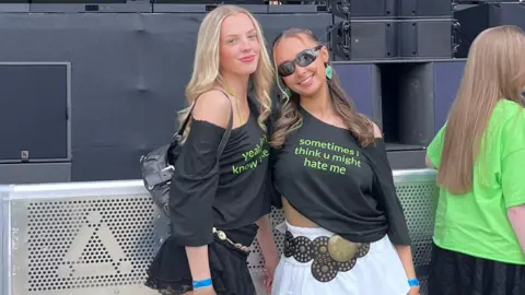 Anna Lawther Two girls in black tops in front of a stage. The girl on the left has blonde hair and the girl on the right has brown hair. The girl on the left wears a black skirt and the girl on the rihgt wears a white skirt. The girl on the right wears sunglasses.