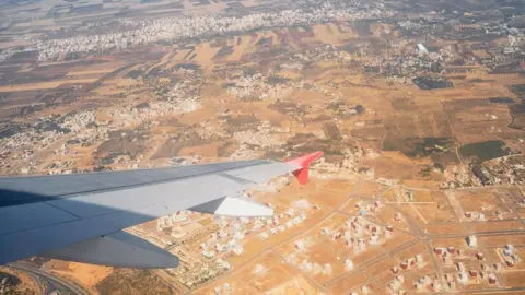 Michael Hall An aerial view of Morocco out of a plane window. The landscape is sandy coloured with sections of green. The plane wing is in shot.
