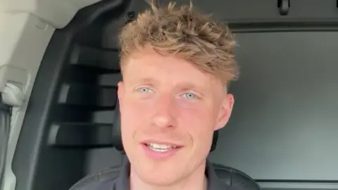 A young man with short hair sitting in a van.