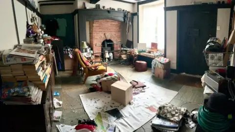 Mark Cargill A room filled with clutter including books, furniture and boxes. It is the inside of a black and white timbered house and you can see a brick fireplace in the background. On the floor is a white sheet with a red stain.