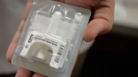 A hand holding a packet of medication up to the camera. In the packet is a nasal spray, with a generic non-brand label reading 'Naloxone HCI Nasal Spray".