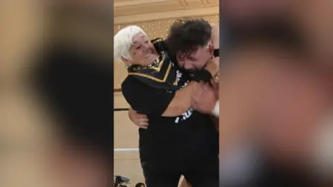 A woman with short, white hair has a man in a headlock