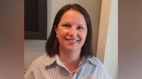 A woman wearing a striped shirt is smiling at the camera. She has brown hair and is standing indoors with what appears to be a television screen attached to a wall behind her.