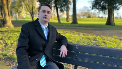 A man with black hair, swept to his left side, sits on a bench in a park. He wears a black coat, partly buttoned, over a light shirt and a blue striped tie with dark trousers. The wooden bench sits in front of a grasses area with some trees in the distance.