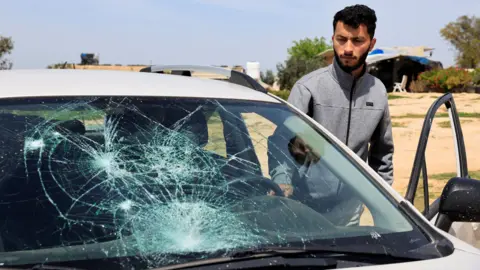 Basel Adra looks at a grey car with its window smashed.