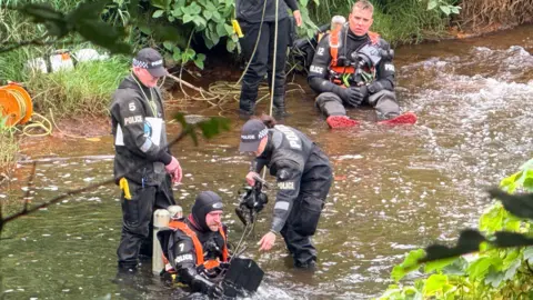 Divers in the River Irwell - a man in a police hat stands up - with a man sitting in the water beneath him with oxygen tanks on his back - a woman in a police hat and outfit holds a camera