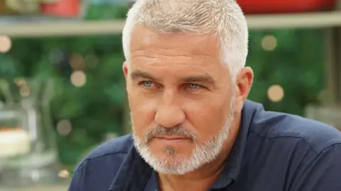 A man with white hair and heard, wearing a dark blue polo shirt. 