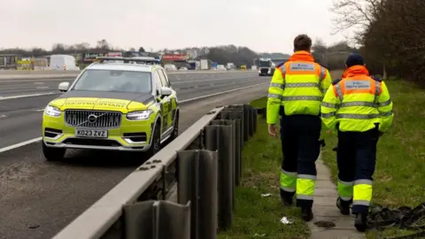 National Highways Two highways officers in high-visibility jackets walking along a grass verge. A National Highways branded car is parked on the side of a motorway.
