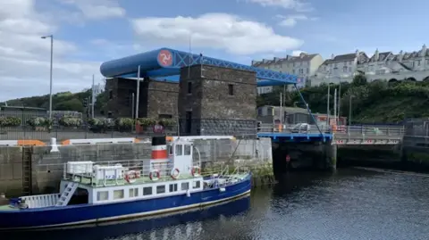 A chug boat in the foreground with the Millennium Bridge behind it, a blue structure with the Manx three legs of man symbol on it, over Douglas Harbour.