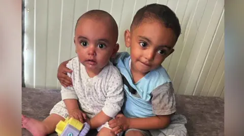 Two year old Mohamed is being hugged by his older brother Noah, they are sat on the floor