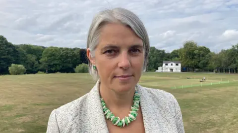 A woman with a silver and white short hairstyle wearing a green and white coloured necklace and grey jacket. There is a white cricket pavilion behind her and a green cricket pitch.