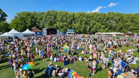 Jason Taylor A general view of Jesus Green in Cambridge during the 2025 Cambridge Pride event. There are hundreds of people in the field and a stage is in the distance.