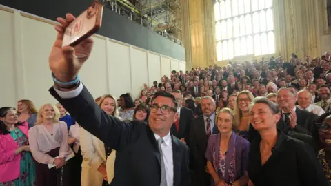 Yui Mok/PA Wire Andy Burnham, with his right arm fully extended, raised above his head and holding a smart phone, taking a selfie alongside a couple of hundred colleagues inside Westminster Hall. He is wearing a dark suit, white shirt and silver tie, and dark rimmed glasses. He and the fellow MPs behind him are smiling broadly. Behind them in the distance is a huge brightly light window.