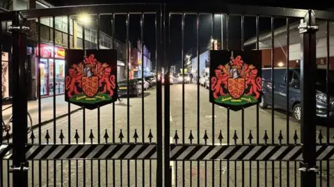 A large black bi-fold set of gates with coats of arms on each one on a commercial street in Gloucester city centre. Parked cars can be seen on the road, which has fast food venues on.