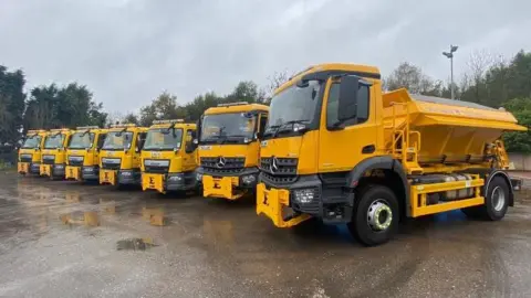 Stoke-on-Trent City Council A row of yellow gritting lorries with grit piled up on their loaders at the back. They stand on grey tarmac in front of green trees.