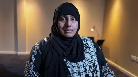 Marium Abbas, wearing a hijab and a white and black patterned top, staring into the camera. She is in a room with a desk behind her, which has a light on top.