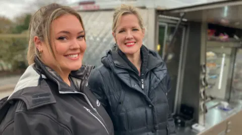 Two women in a black jackets are looking towards the camera, smiling. Behind them is a catering vehicle, with metal shelves and an oven area