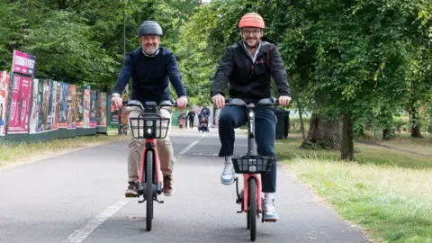 City of Edinburgh Council Transport and Environment Convener,Stephen Jenkinson (left) wearing a dark jumper and light trousers and Voi UK General Manager, James Bolton (right) wearing dark clothing test out the new e-bikes at the launch of Edinburgh's trial cycle hire scheme.