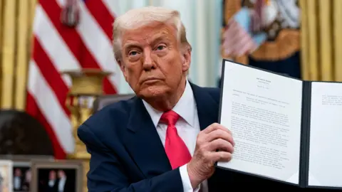 EPA Donald Trump wears a blue suit, white shirt and red tie. He has grey hair. He holds the signed proclamation.