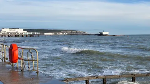 Sparrow A seascape with several waves breaking against the sea wall. There is a pier with two white buildings and a cliff head in the distance. The sky is blue and the sun is shining.