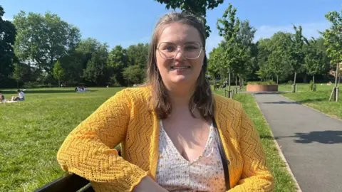 A woman with blonde hair, glases and a yellow cardigan and flowery white top.