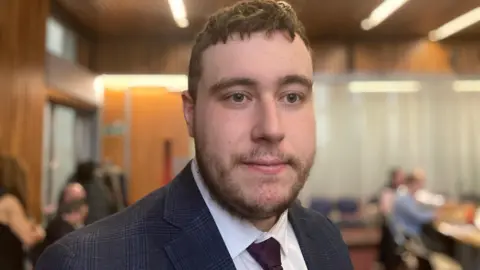 Laura Coffey/BBC Jack Goncalvez in a navy suit and maroon tie,