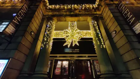 Hebden Bridge Picture House The wooden entrance doors to the building, between two large concrete columns. The columns are covered in gold and white coloured Christmas lighting, and a large lit-up decoration in the shape of a bow and ribbon is over the doors.