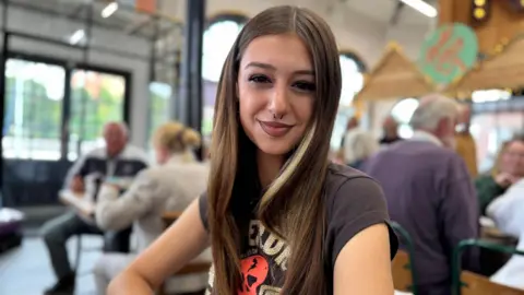 Layla has long straight dark hair and is wearing a dark t-shirt. She's sitting in a busy cafe smiling.
