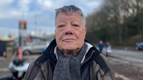 Elizabeth Baines / BBC A man stands in a brown coat. He wears a grey scarf and behind him a picket line is blurred.