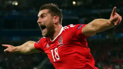 Sam Vokes in the red jersey of Wales shouts in celebration as he runs with arms outstretched after scoring a goal
