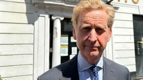 Ben Parker/BBC John Howard wearing a navy suit and blue spotted tie, standing in front of the Great White Horse Hotel. The building is white with wood panels.