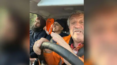 Marcus Lapsa Bill Mato, Kully Kaur Deol and Marcus Lapsa are smiling in their van driving down to Pontypridd. They are wearing blue jackets and Marcus Lapsa is wearing an orange hi-vis jacket. Mr Lapsa is driving.