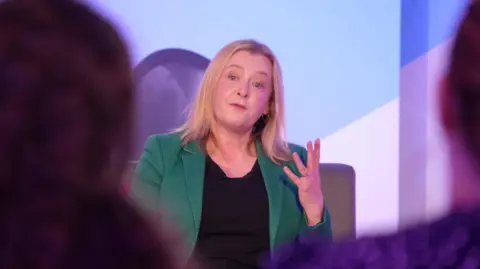 Jayne Bryant gesticulates with her hand, as she sits on a chair on the studio set. The backs of the heads of blurred audience members are either side of her nearer the camera. She is wearing a green jacket over a black top.