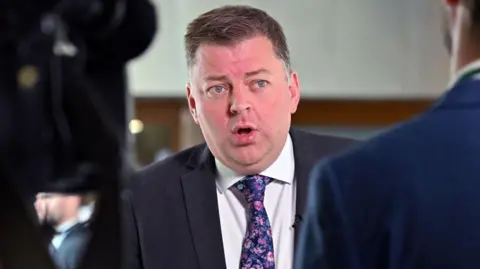 Getty Images Colin Smyth, a man with spiky brown hair mid speech while wearing a suit, white shirt and blue flowery tie